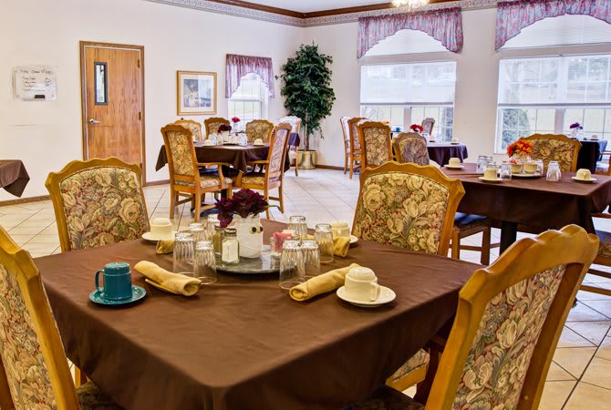 A cozy dining room with elegantly set tables and upholstered chairs, featuring large windows and decorative elements.