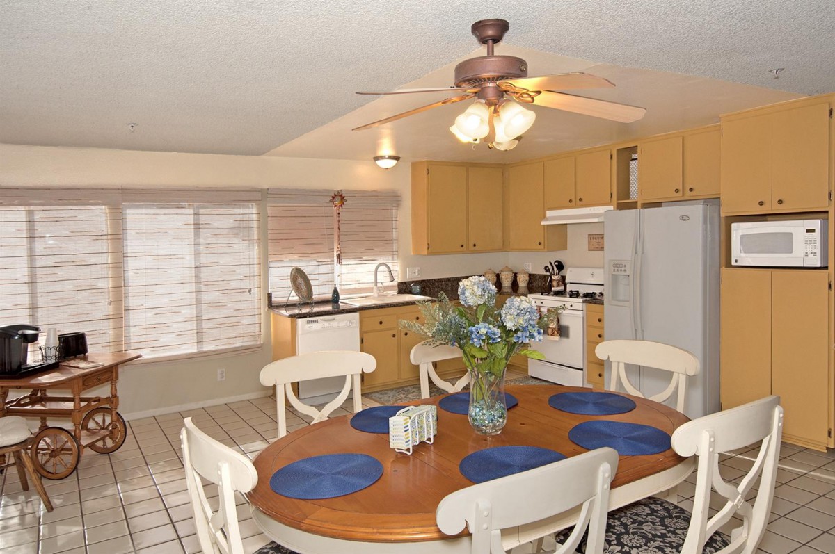 A cozy dining area with a wooden table set for four, featuring blue placemats and a centerpiece of flowers, adjacent to a kitchen equipped with modern appliances.
