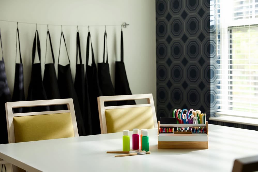 A well-organized art room with aprons hanging on the wall, and a table set with craft supplies including paint bottles and scissors.