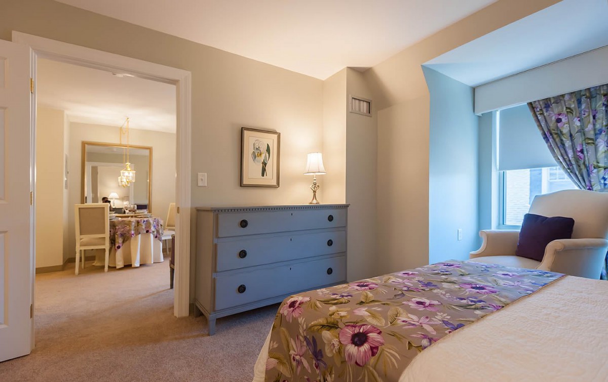 A cozy bedroom with a floral bedspread and a blue dresser, softly illuminated by natural light.