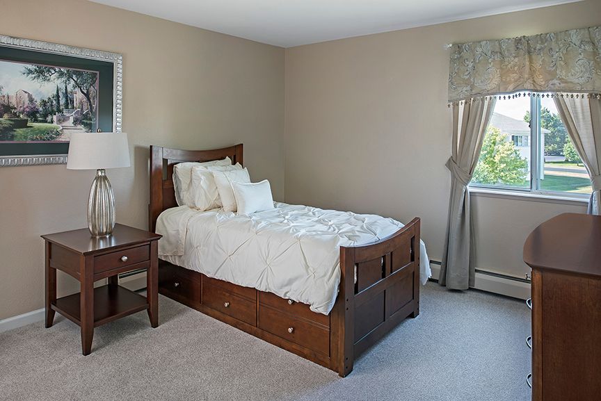 A cozy bedroom featuring a single wooden bed with built-in storage, a nightstand with a lamp, and a window dressed with curtains.