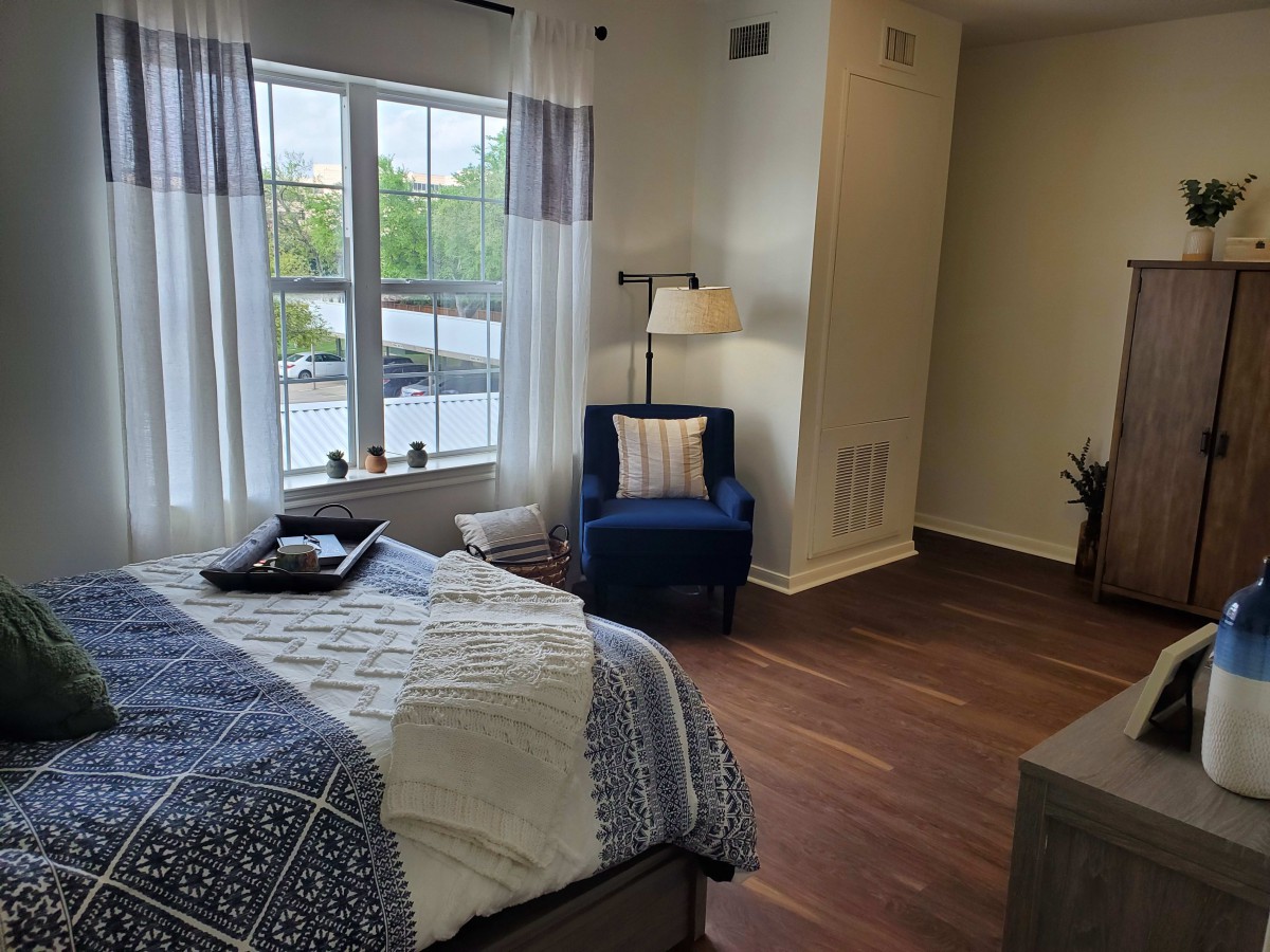 A cozy bedroom with a large window, featuring a bed with patterned bedding, a comfortable chair, and a standing lamp.