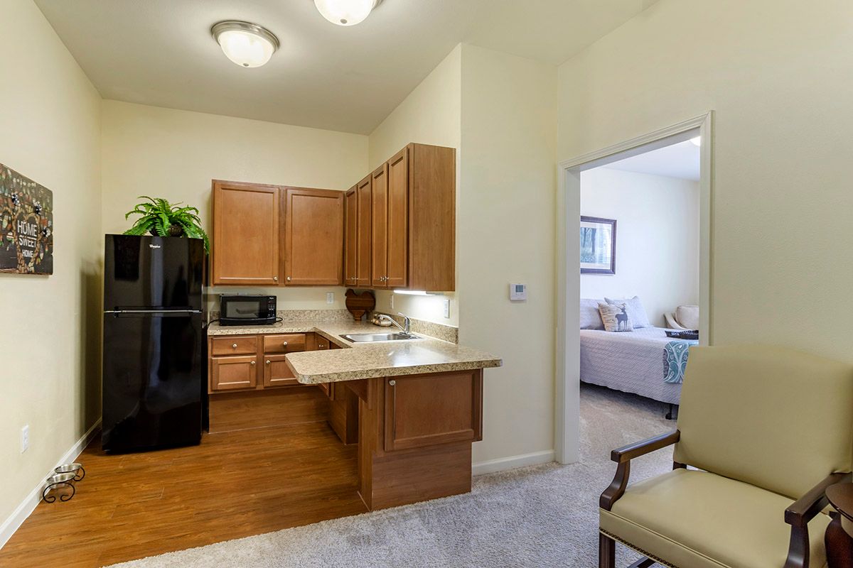 A cozy apartment kitchen with wooden cabinets and black appliances, leading into a tastefully decorated living room.