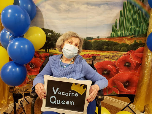 An elderly woman proudly holding a sign that reads 'Vaccine Queen,' celebrating her vaccination with balloons in the background.