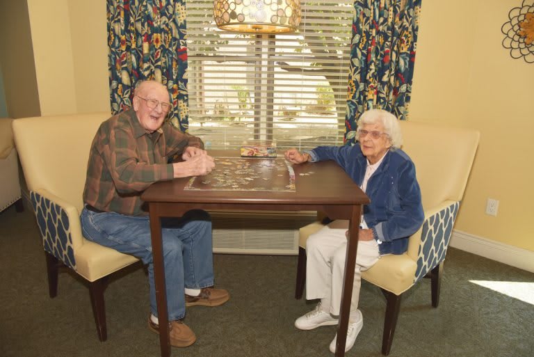 Two elderly individuals sitting at a table in a cozy room with patterned curtains.