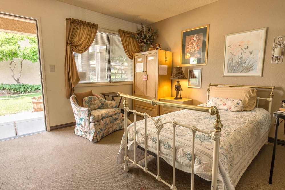 A cozy vintage-style bedroom with an ornate bed, armchair, cabinet, and window with curtains.