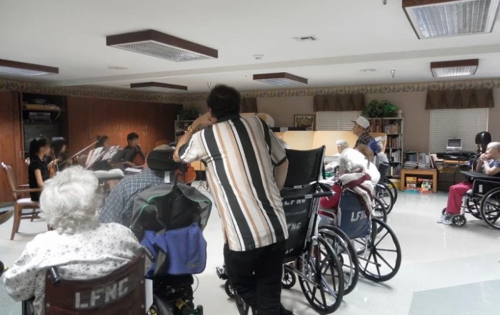 A caregiver assists elderly individuals in wheelchairs at a nursing home's community room.