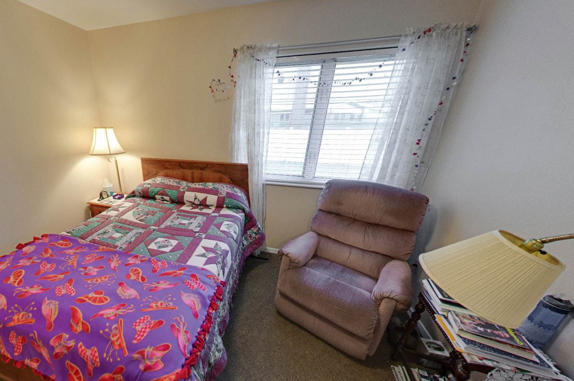 A cozy bedroom featuring a bed with a colorful quilt, a reclining chair, a window with curtains, and a lamp.