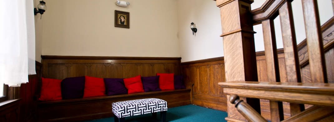A cozy interior room with wooden paneling, bench seating adorned with colorful cushions, and a patterned ottoman. A portrait and wall sconce decorate the walls.