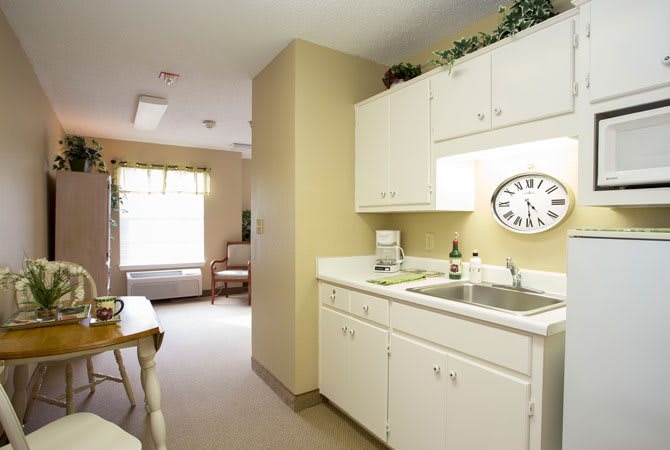 A small kitchen area with white cabinets and a sink, featuring a wall clock and natural light coming through the window.