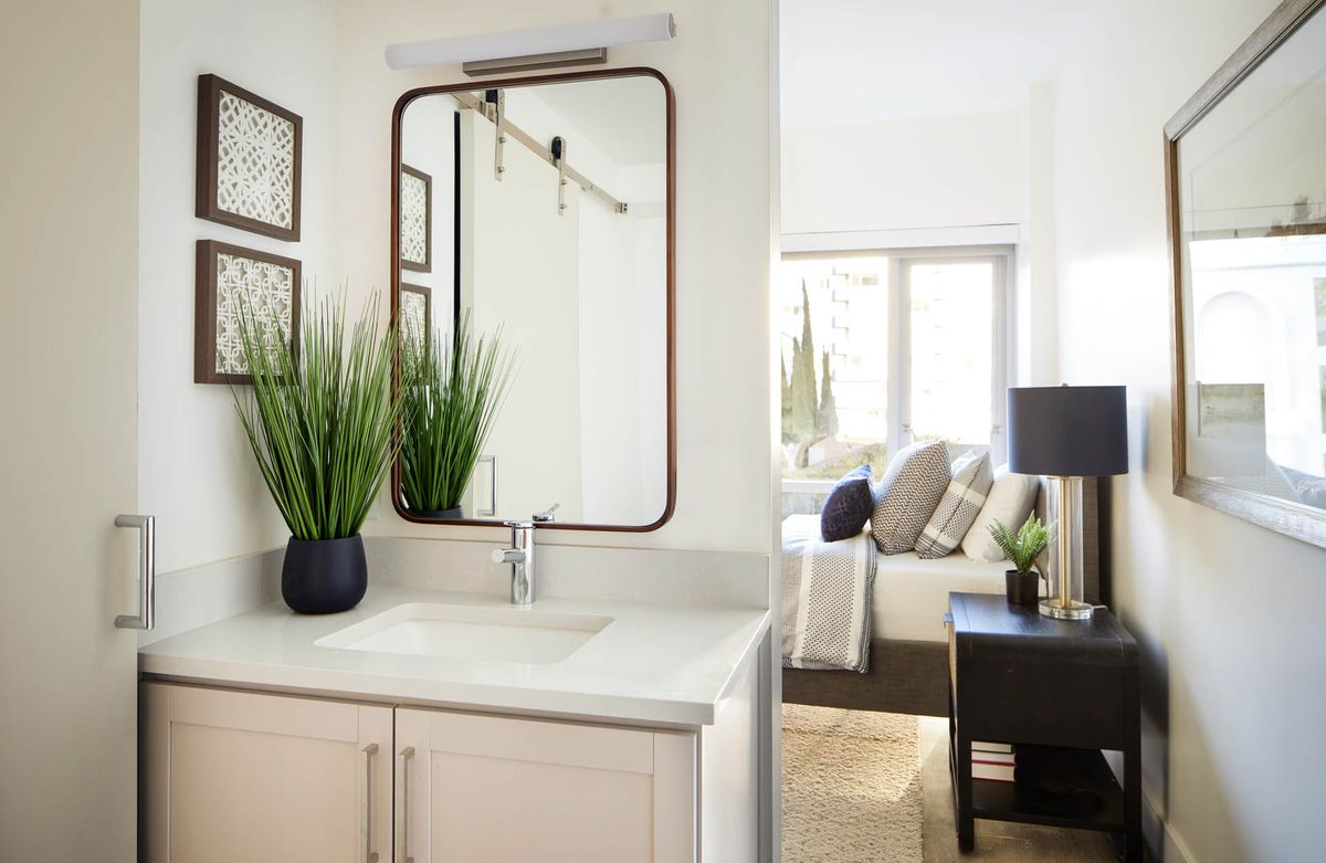 A modern bathroom with a large mirror and potted plants, adjacent to a cozy bedroom.
