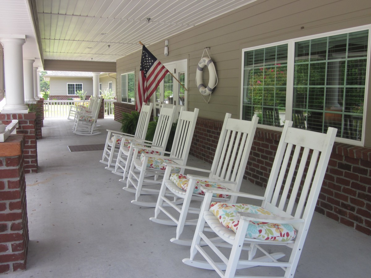 A row of white rocking chairs lined up on a spacious porch, adorned with an American flag.