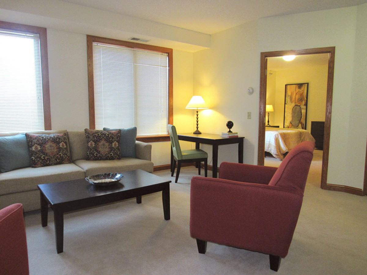 A cozy living room featuring a red armchair, a sofa with patterned cushions, a coffee table, and a warm-lit lamp.
