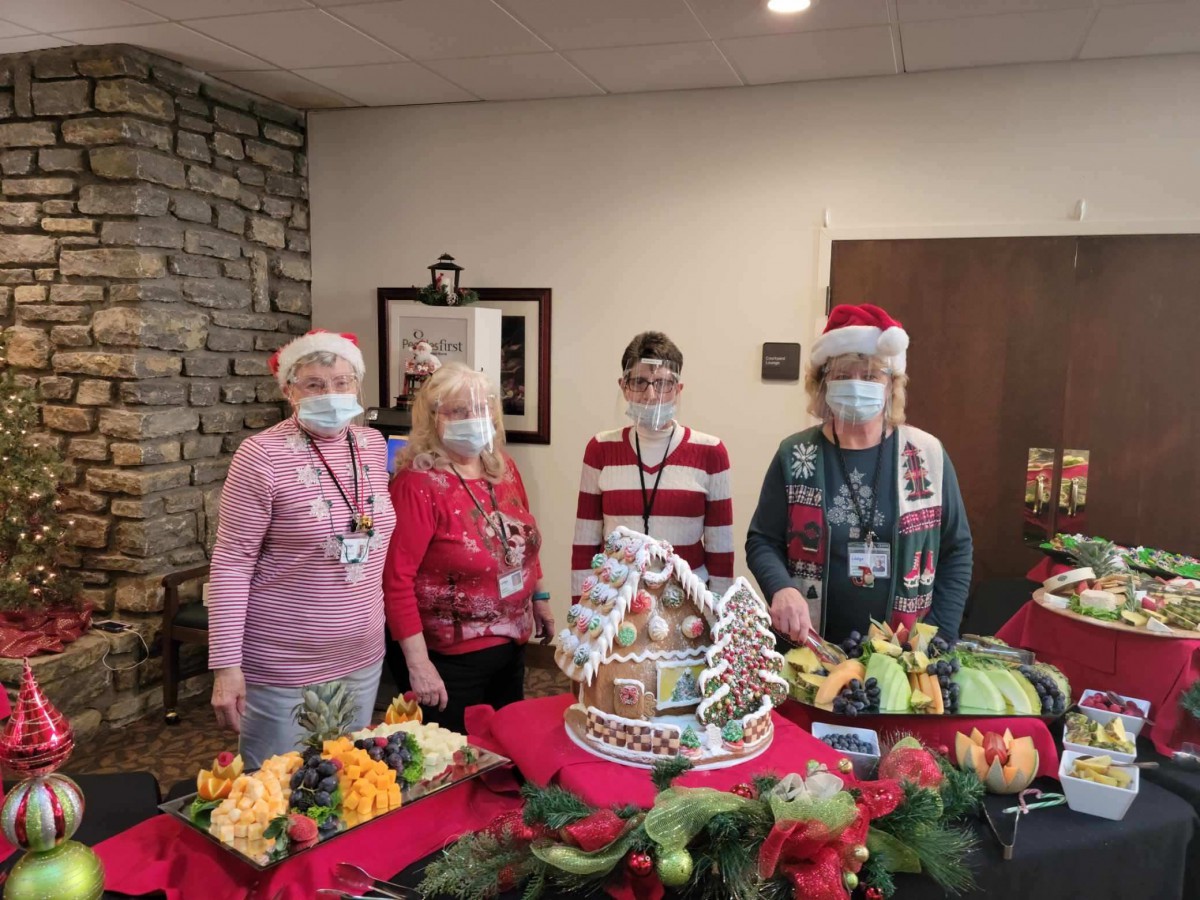Four people in festive attire stand behind a table with a decorated gingerbread house and holiday decorations.