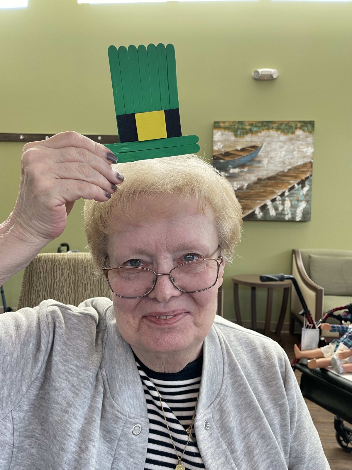 An elderly woman playfully holds a green paper hat above her head while smiling indoors.