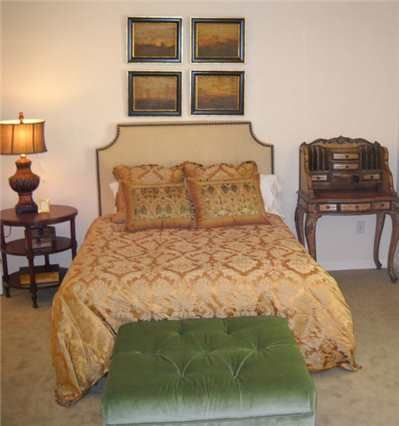 A cozy bedroom featuring a bed with decorative pillows, a side table with a lamp, artwork on the wall, and a small desk.