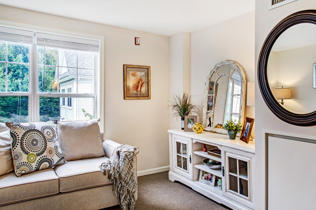 A cozy living room featuring an armchair beside a window and a decorative white cabinet with a mirror.