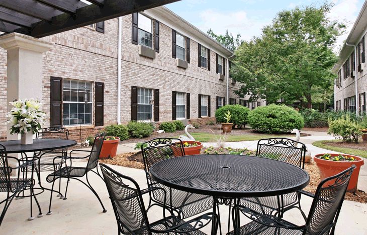 A tranquil outdoor patio area with tables and chairs, surrounded by landscaped greenery in front of a residential building.