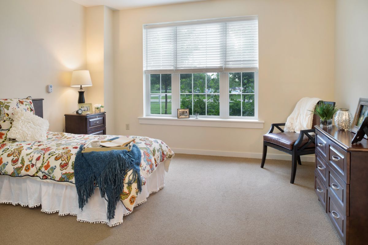 A cozy bedroom with a large window, neatly arranged furniture including a bed, lamp, and chair.