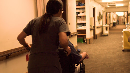 A nurse assists a patient in a wheelchair down a hospital corridor.