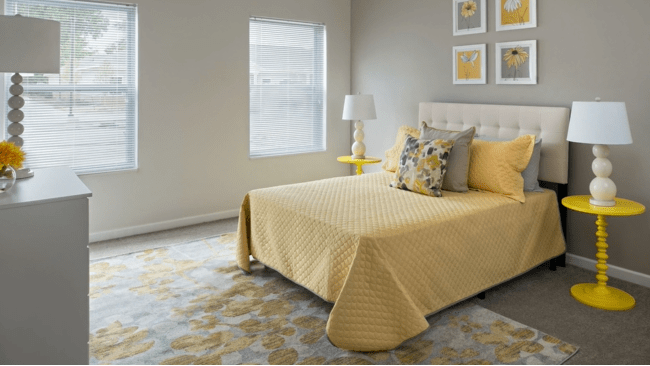 A modern bedroom with yellow bedding and decorative pillows, featuring a lamp and framed artwork on the neutral-colored walls.