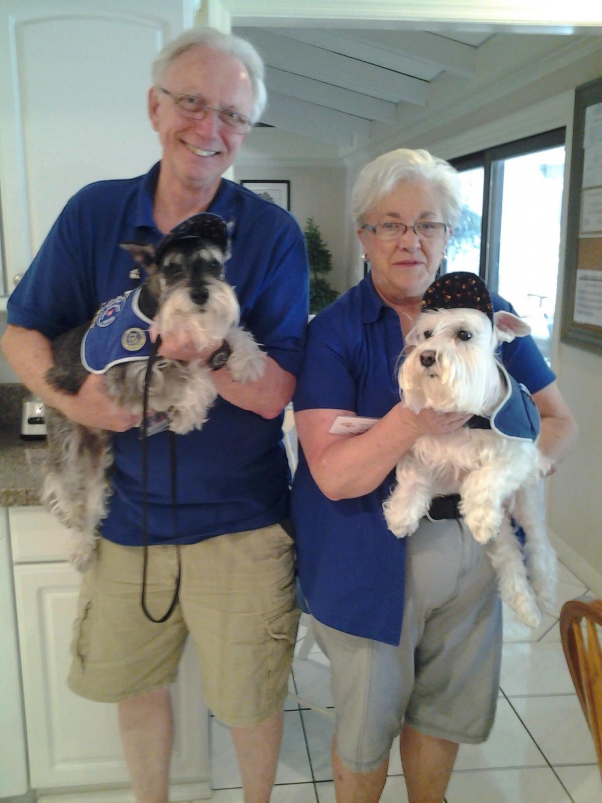 Two people smiling and holding their dogs dressed in matching outfits indoors.