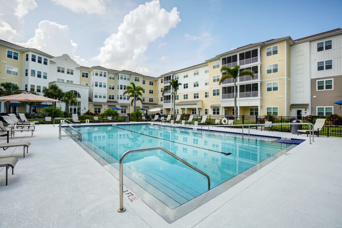 A modern apartment complex featuring an outdoor swimming pool surrounded by palm trees on a sunny day.