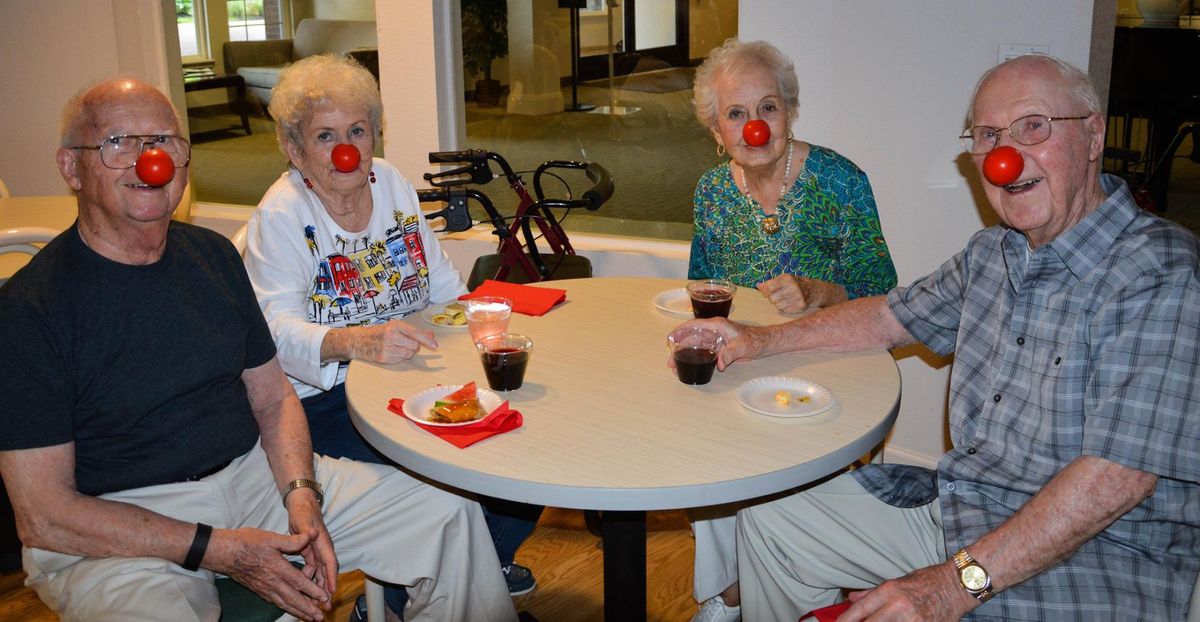 A group of elderly women wearing red noses gather around a table, enjoying each other's company.