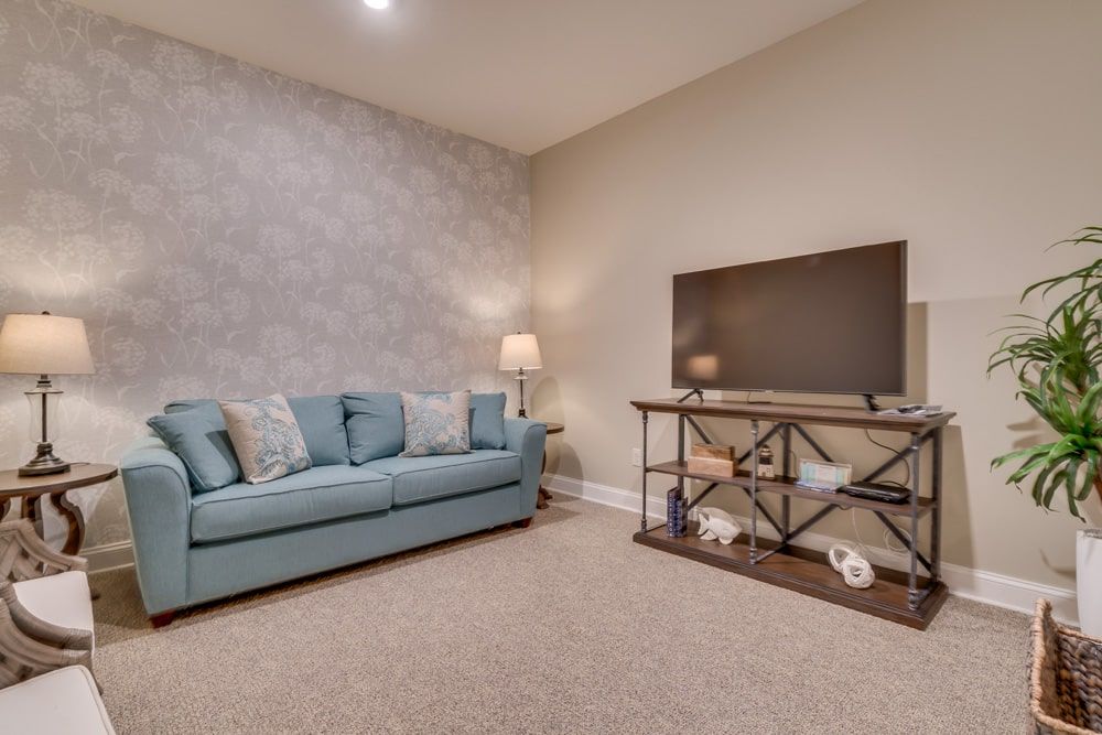 A cozy living room with a blue sofa, television, and lamp, featuring neutral colored walls and carpeted flooring.