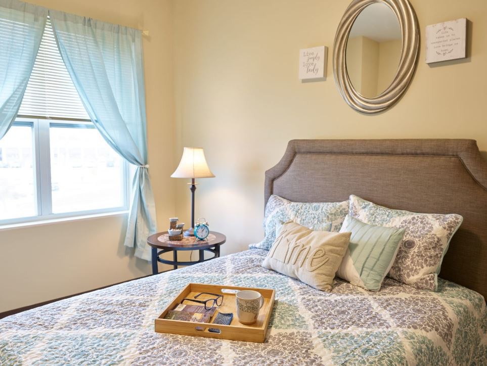 A cozy bedroom with a neatly made bed, decorative pillows, and a bedside table with a lamp. The room features a mirror on the wall and soft curtains framing the window.