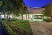 A view of a building exterior at night, featuring a well-lit entrance, surrounding garden, and walkway.