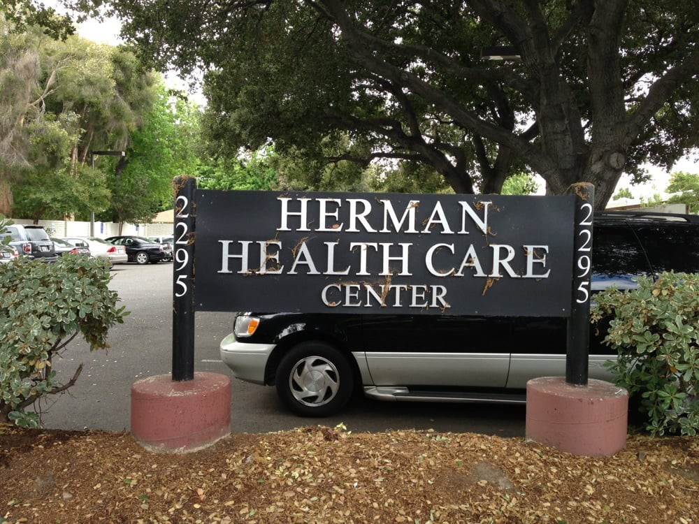 A sign for Herman Health Care Center situated in a parking area with trees and a parked car.
