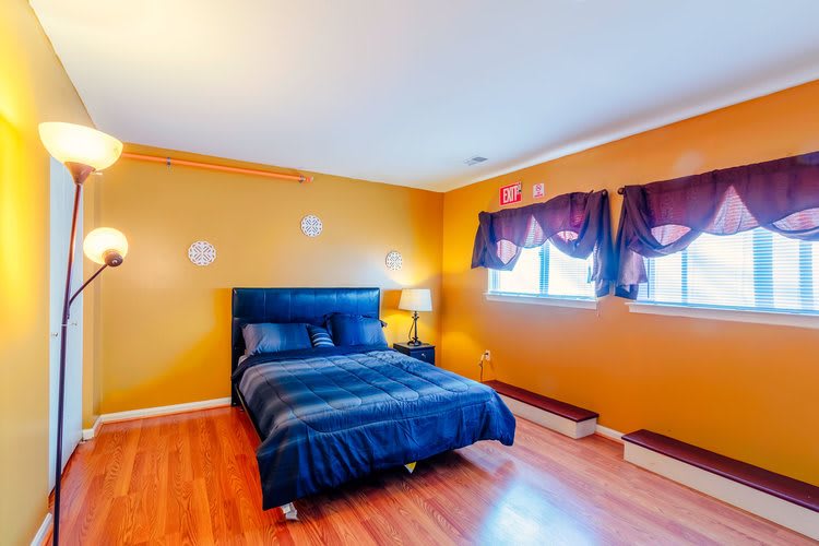 A cozy bedroom with vibrant orange walls and blue bedding, featuring wooden flooring and windows adorned with curtains.