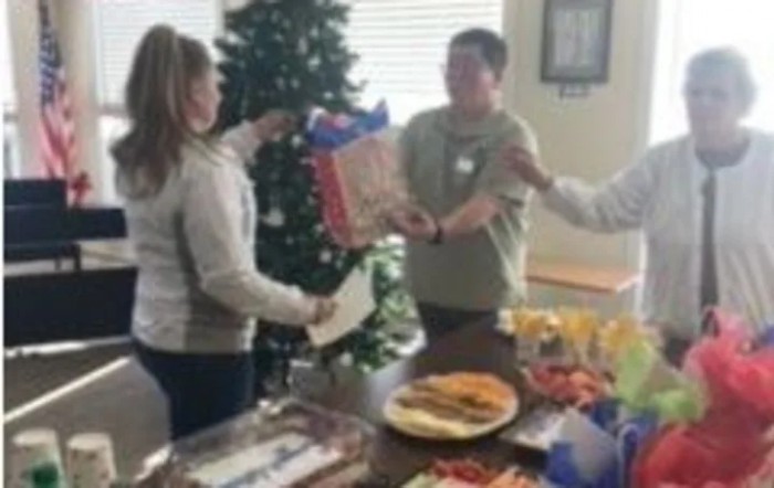 People exchanging gifts at a holiday party with food on the table and a decorated Christmas tree in the background.