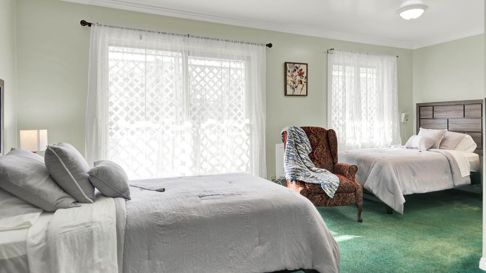 A cozy bedroom with large windows, a comfortable bed, and an armchair draped with a blanket.