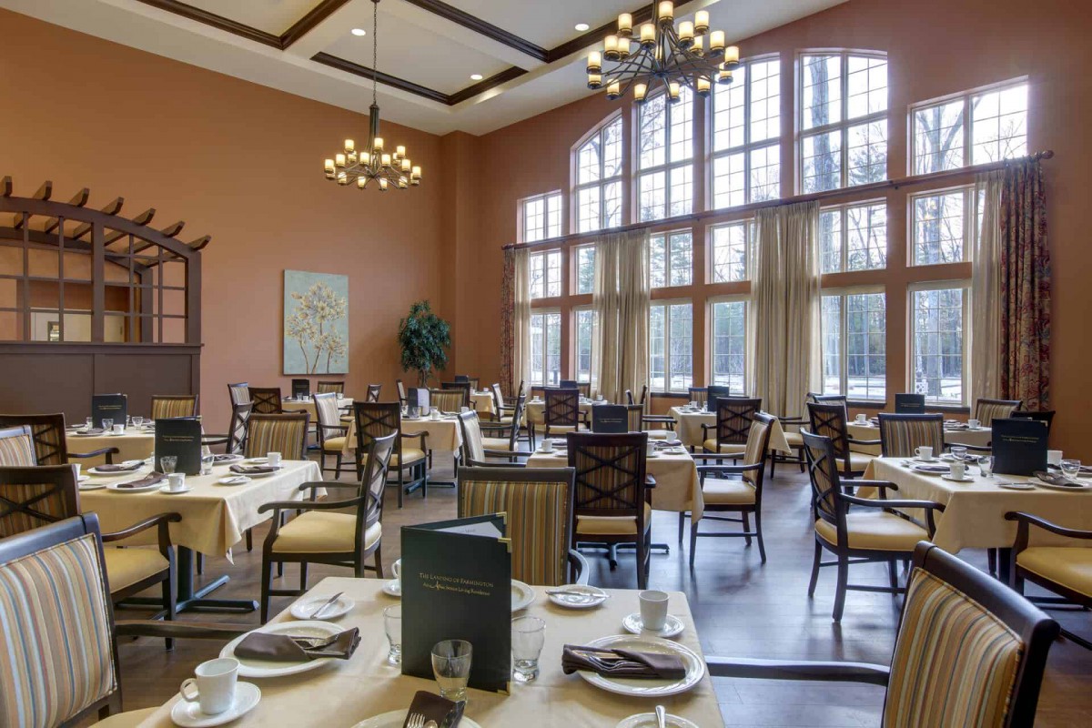 An elegant dining room with large windows and chandeliers, featuring neatly arranged tables.
