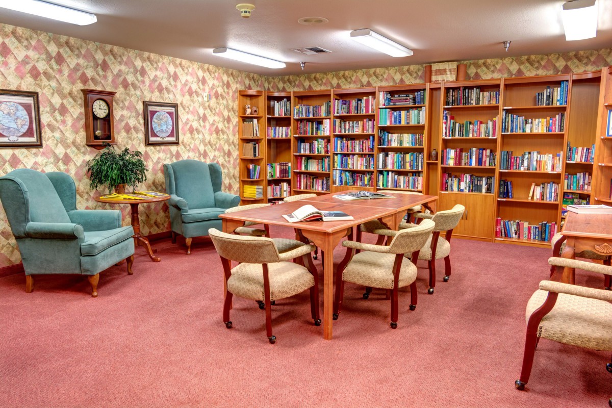 A cozy reading room with bookshelves, a central table surrounded by chairs, and comfortable armchairs.