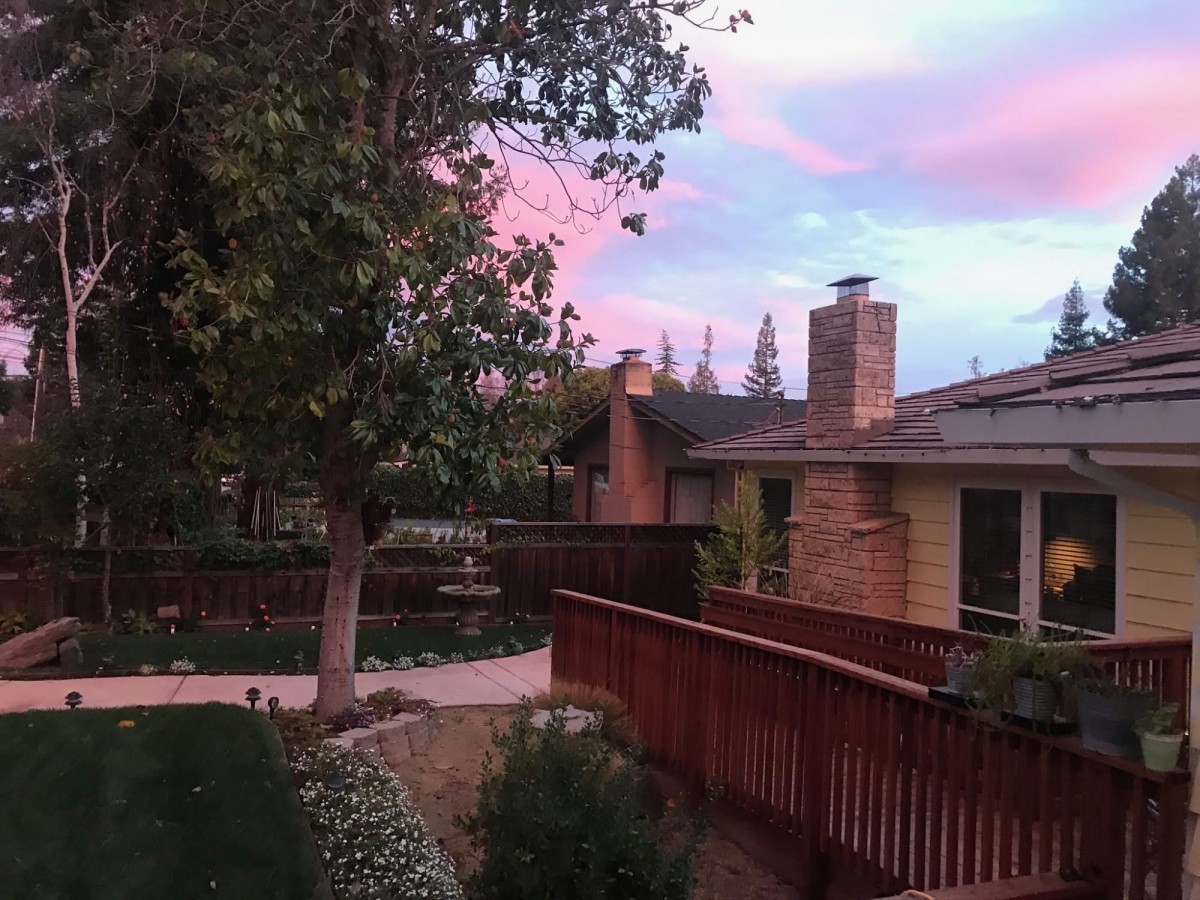 A serene backyard view at sunset, featuring a house surrounded by trees and a wooden fence under a colorful sky.