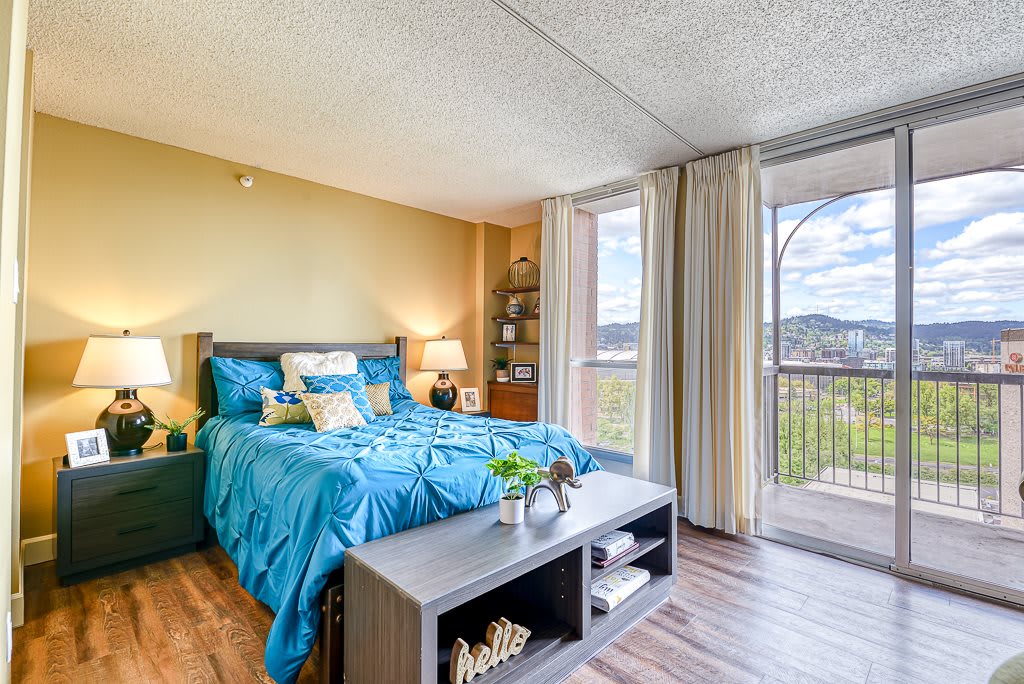 A modern bedroom with blue bedding and natural light streaming through large windows, offering a view of the balcony.
