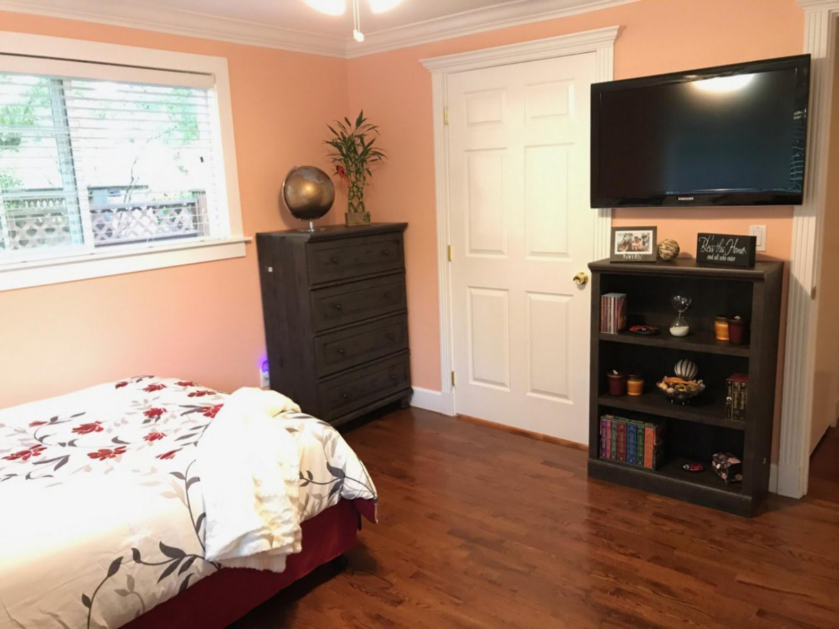 A cozy bedroom with warm peach walls, featuring a bed with floral bedding, a tall dresser, and a wall-mounted television above a small bookshelf.