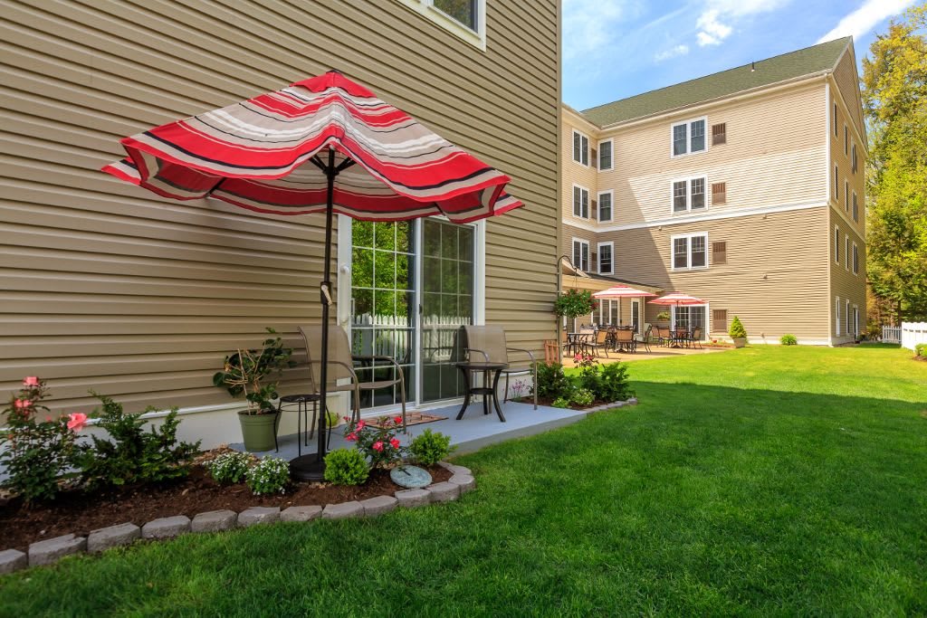 A sunny backyard patio with a red and white umbrella, surrounded by a well-maintained lawn and garden in an apartment complex.