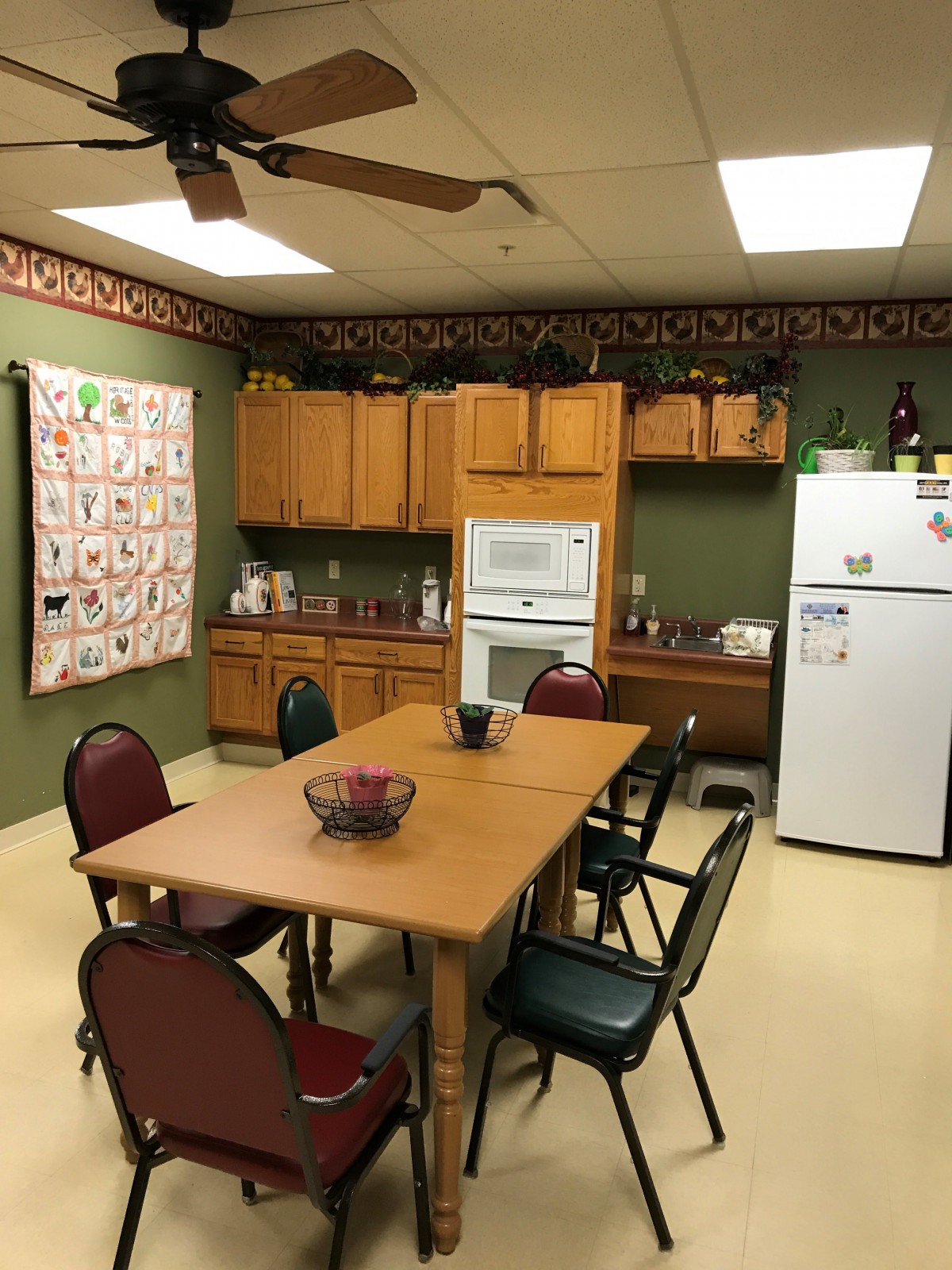 A cozy kitchen and dining area featuring a table with chairs, wooden cabinets, a refrigerator, and a microwave. A quilt decorates one of the walls.