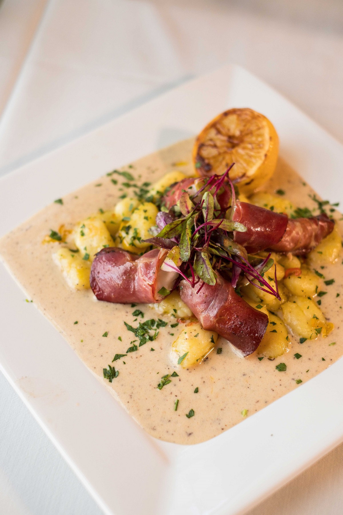 A gourmet dish featuring sausages with potatoes, drizzled with sauce and garnished with fresh herbs and a grilled lemon half.