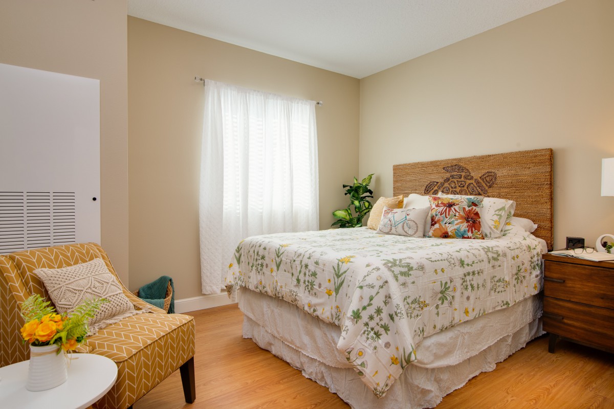 A cozy bedroom with a floral bedspread, wooden flooring, and soft natural light coming through the window.
