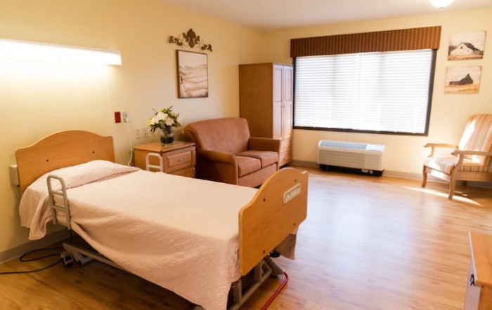 A clean and well-lit hospital room with a single bed, a comfortable chair, and a large window.