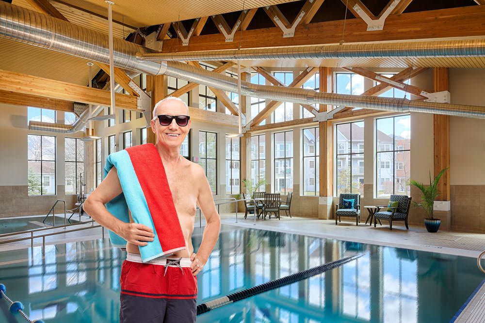 A person wearing sunglasses and a towel stands confidently by an indoor swimming pool.