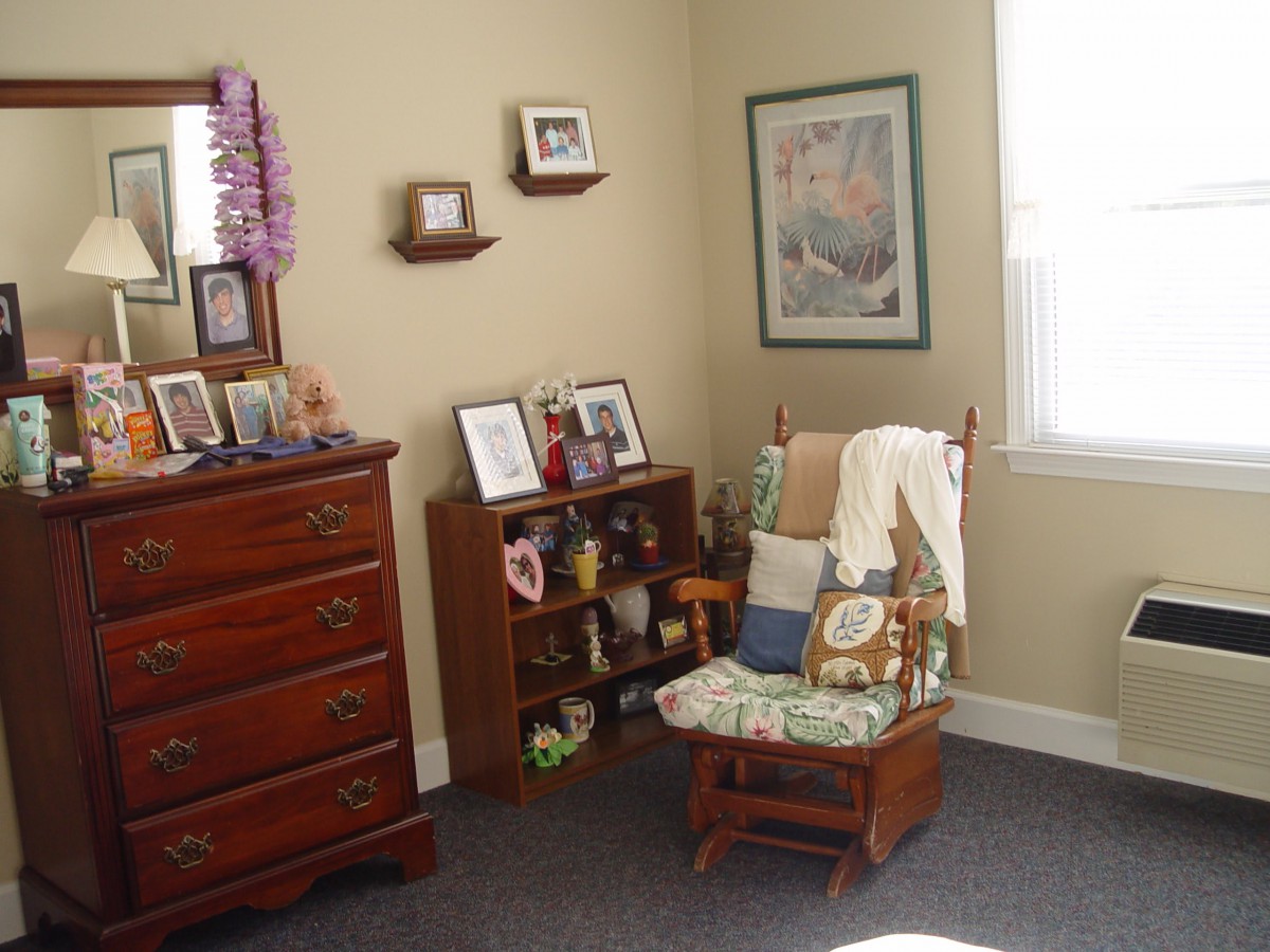 A cozy corner of a room featuring a rocking chair, small dresser, and shelves adorned with photographs and decorative items.