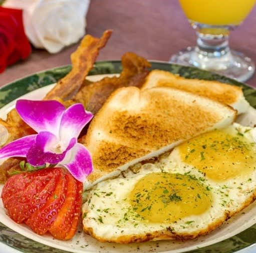 A breakfast plate with sunny-side-up eggs, toast, bacon, and a strawberry slice, garnished with an orchid flower, accompanied by a glass of orange juice.