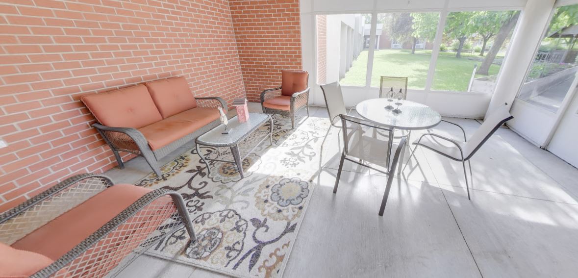 A cozy patio area with comfortable seating, a glass table, and decorative rugs, illuminated by natural sunlight.
