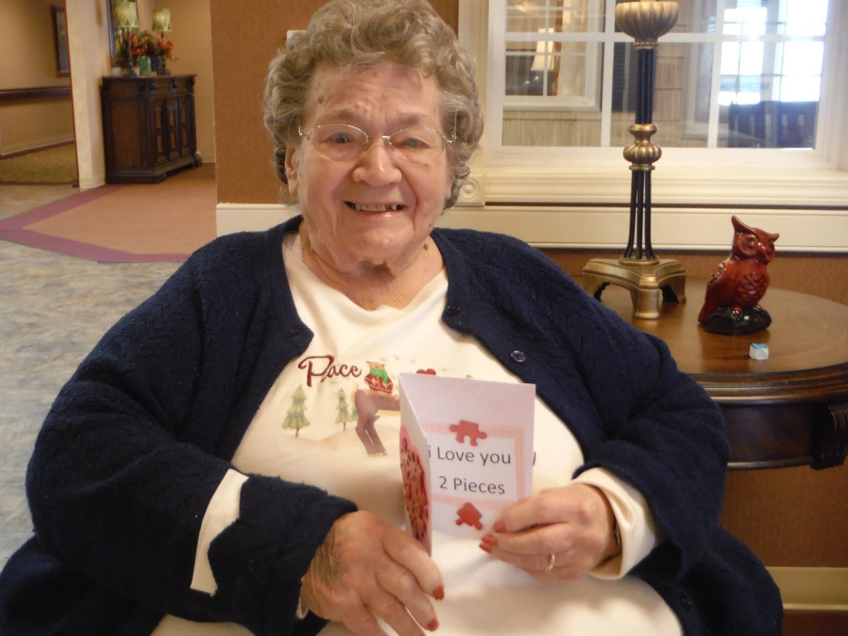 An elderly woman sitting indoors, smiling and holding a book.